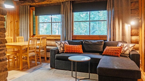 Un interior acogedor de cabaña con una mesa de comedor de madera, sillas, un sofá con cojines y ventanas amplias que ofrecen vistas al bosque.
