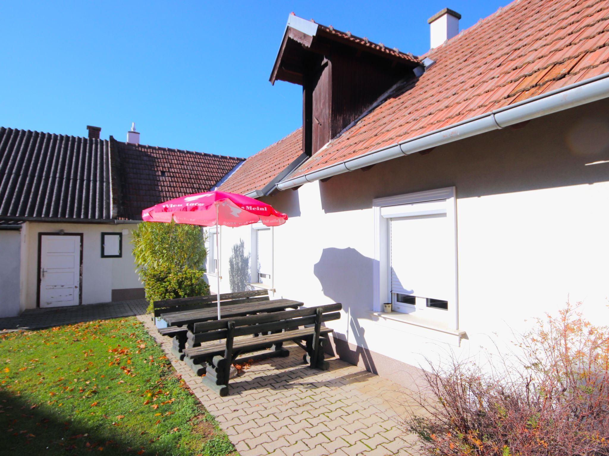 Een gebouw met een pannendak, een witte gevel en een deur. Buiten staan bankjes en er is een roze parasol.
