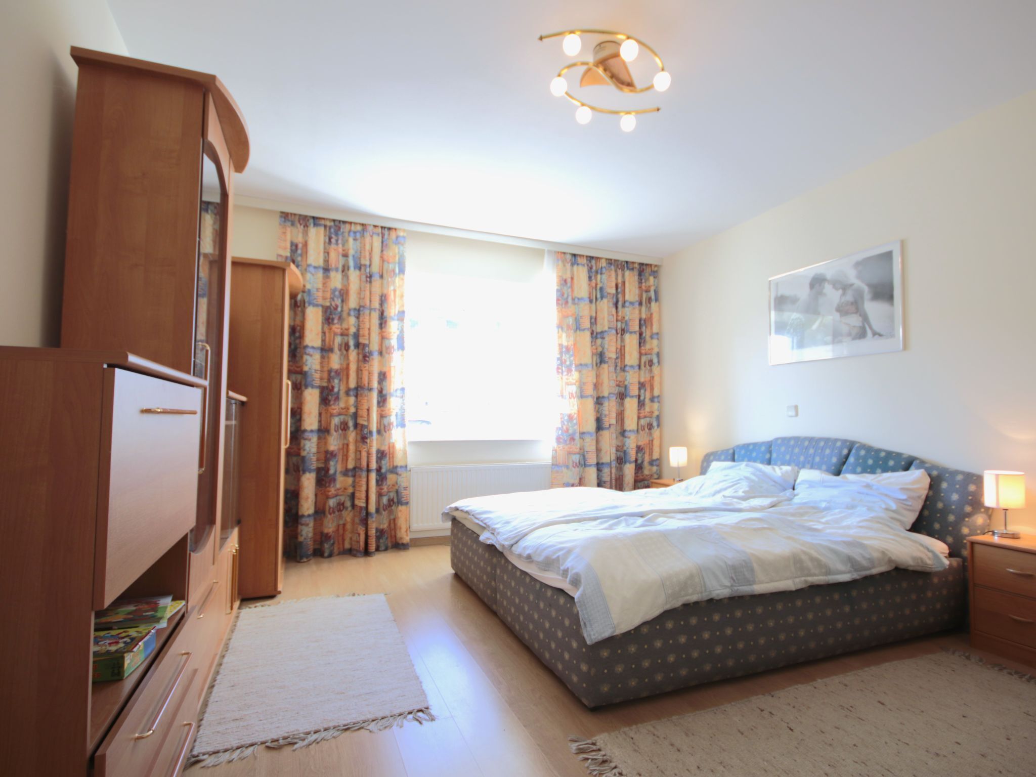 A bedroom with a bed, a wardrobe, a window with curtains, a bedside lamp, and a ceiling light fixture.