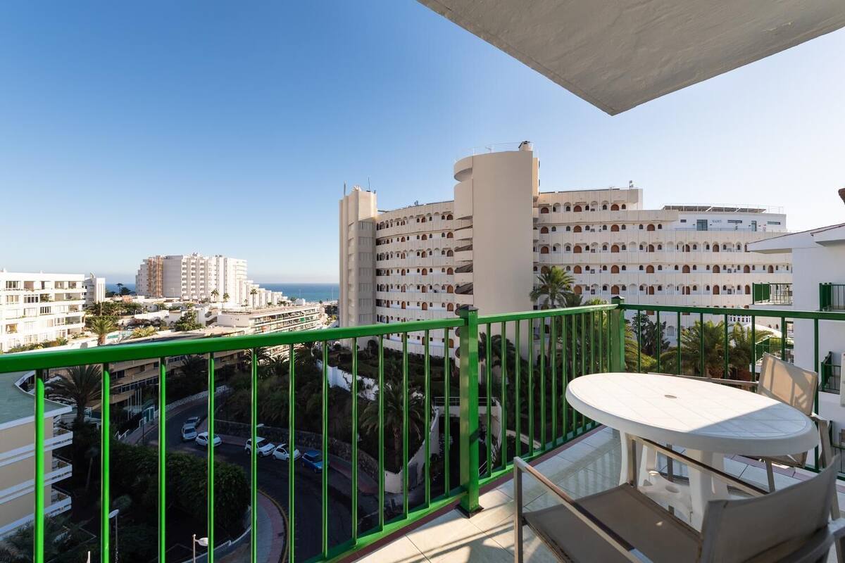 A balcony with a green railing overlooking a coastal cityscape, featuring a dining area with a table and chairs.