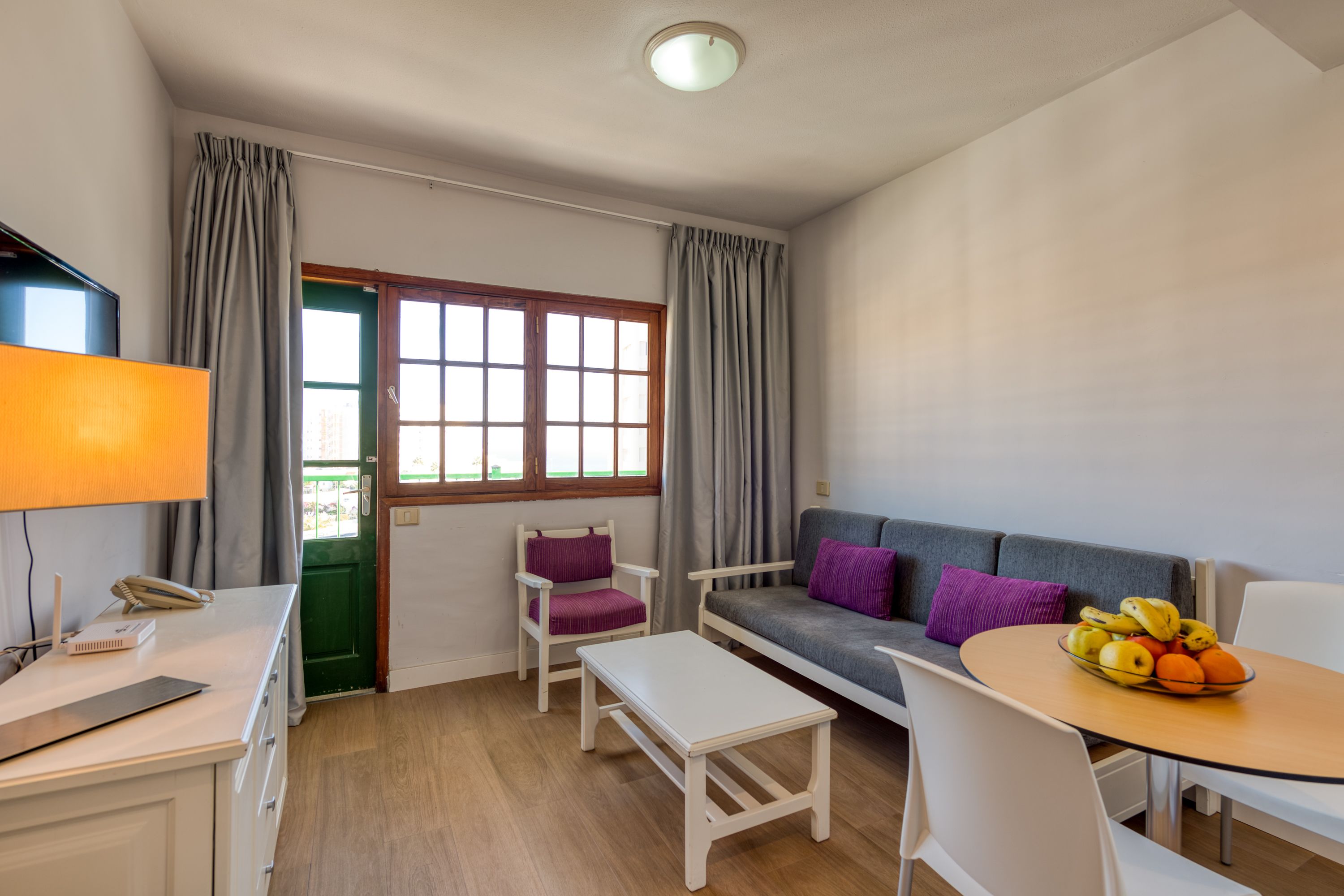 A modern living room with a sofa, a dining table with fruit, a small white table, a chair, a lamp, and a window with curtains.