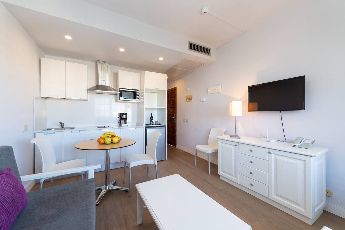 A modern kitchen with white cabinets, a microwave, and a coffee maker. There is a dining table with chairs and a bench. A TV is mounted on the wall, and a lamp is on a cabinet.