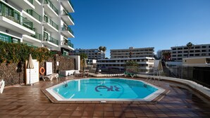 A swimming pool with a tiled deck, surrounded by a fence, and adjacent to a building with multiple balconies and a stone wall. - Apartamentos Don Diego (San Bartolomé de Tirajana)
