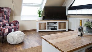 A cozy living room with a TV on a wooden stand, a patterned armchair, and a small round ottoman.