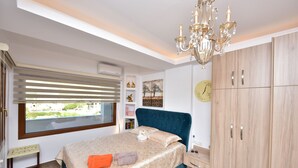 A bedroom with a bed, a chandelier, a wardrobe, a window with blinds, and a view of greenery outside. - Sea Front Panorama Villa Private Pool (Malevizi)