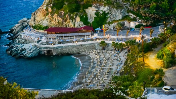 A coastal hotel with a rocky cliff backdrop, a beach with umbrellas, and a dining area with a red-roofed building. - Sea Front Panorama Villa Private Pool (Malevizi)