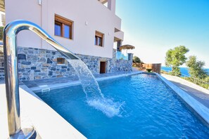 A swimming pool with water flowing from a fountain, a stone wall, and a modern building with windows.