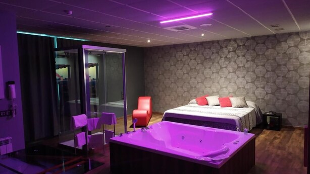 A modern bedroom with a large bed, a glass shower enclosure, a red chair, and a purple jacuzzi tub. - Motel Los Abedules (Oviedo)