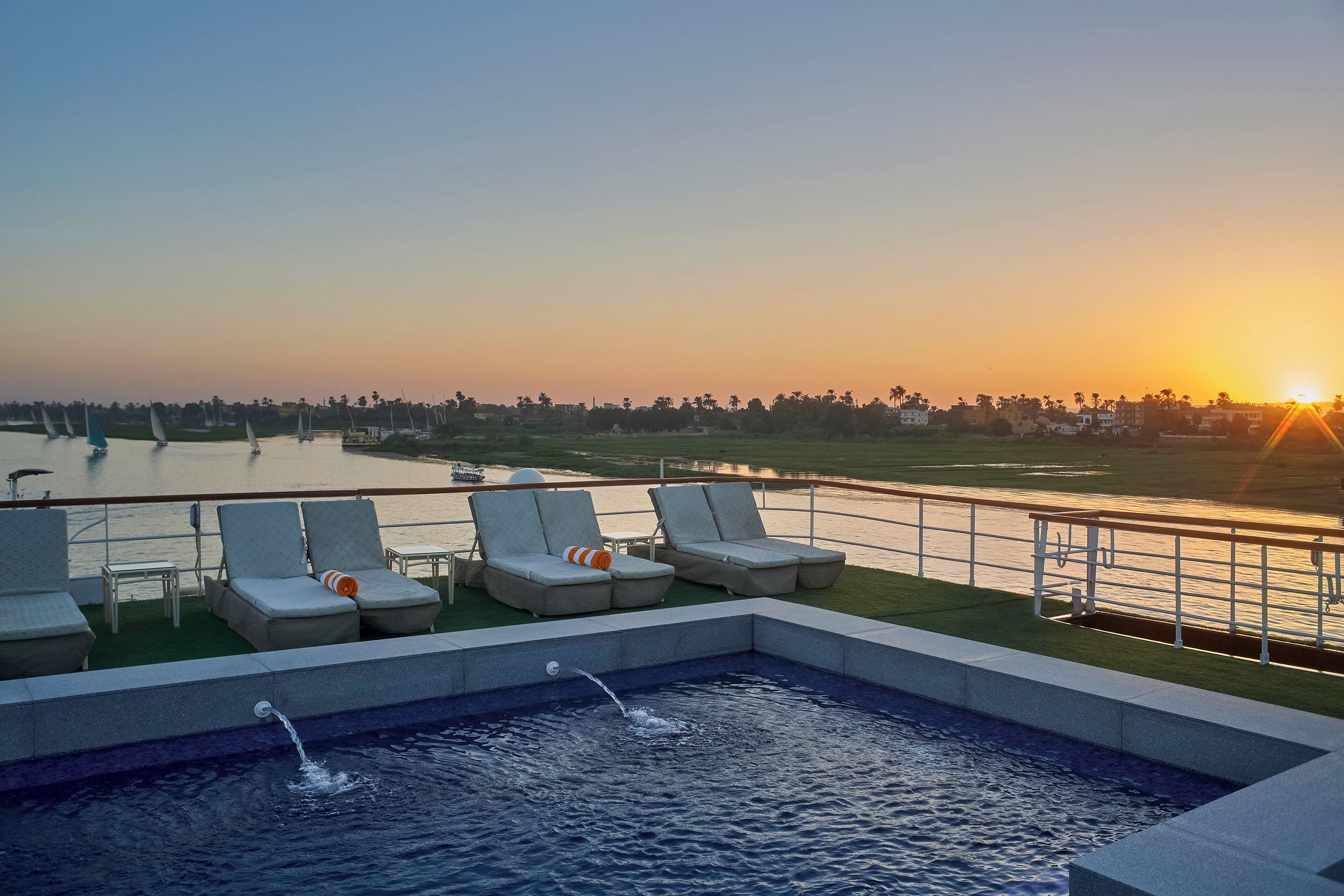 A pool area with lounge chairs and a view of the sunset over a body of water.