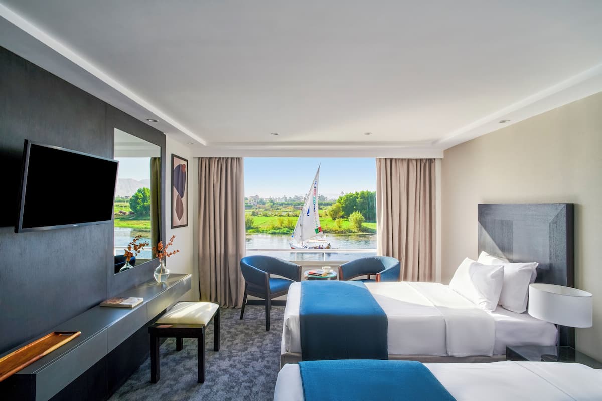 Ett hotellrum med en säng, ett skrivbord, en stol, en TV och utsikt över en flod med en segelbåt.