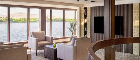 A modern living room with large windows overlooking a body of water, a curved wooden staircase, and a bookshelf with decorative items.