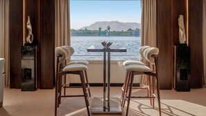 Een kamer met uitzicht op een wateroppervlak, een tafel met stoelen, en twee beelden op sokkels.