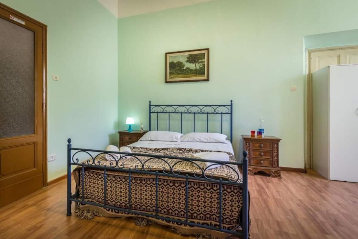 A bedroom with a patterned bed, a nightstand with a lamp, a dresser, and a painting on the wall.