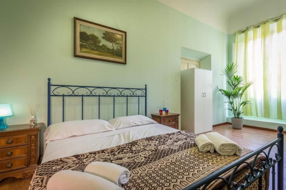 A bedroom with a bed, a nightstand, a dresser, a potted plant, and a window with curtains.
