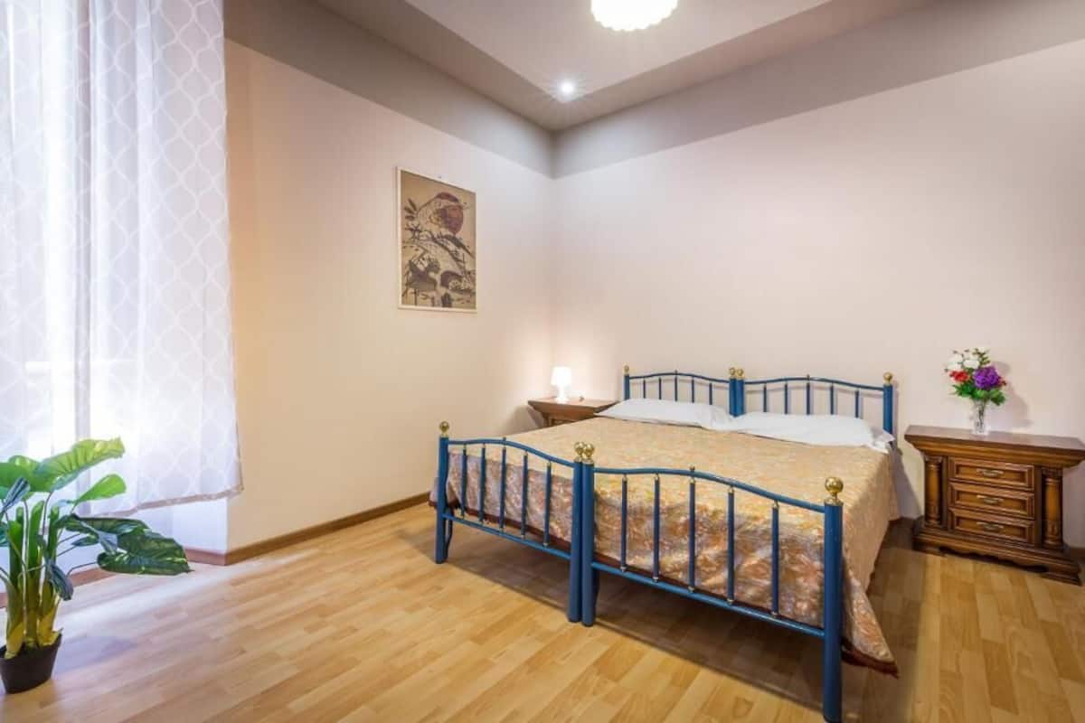 A bedroom with a bed, a nightstand, a dresser, a window with curtains, and a potted plant.