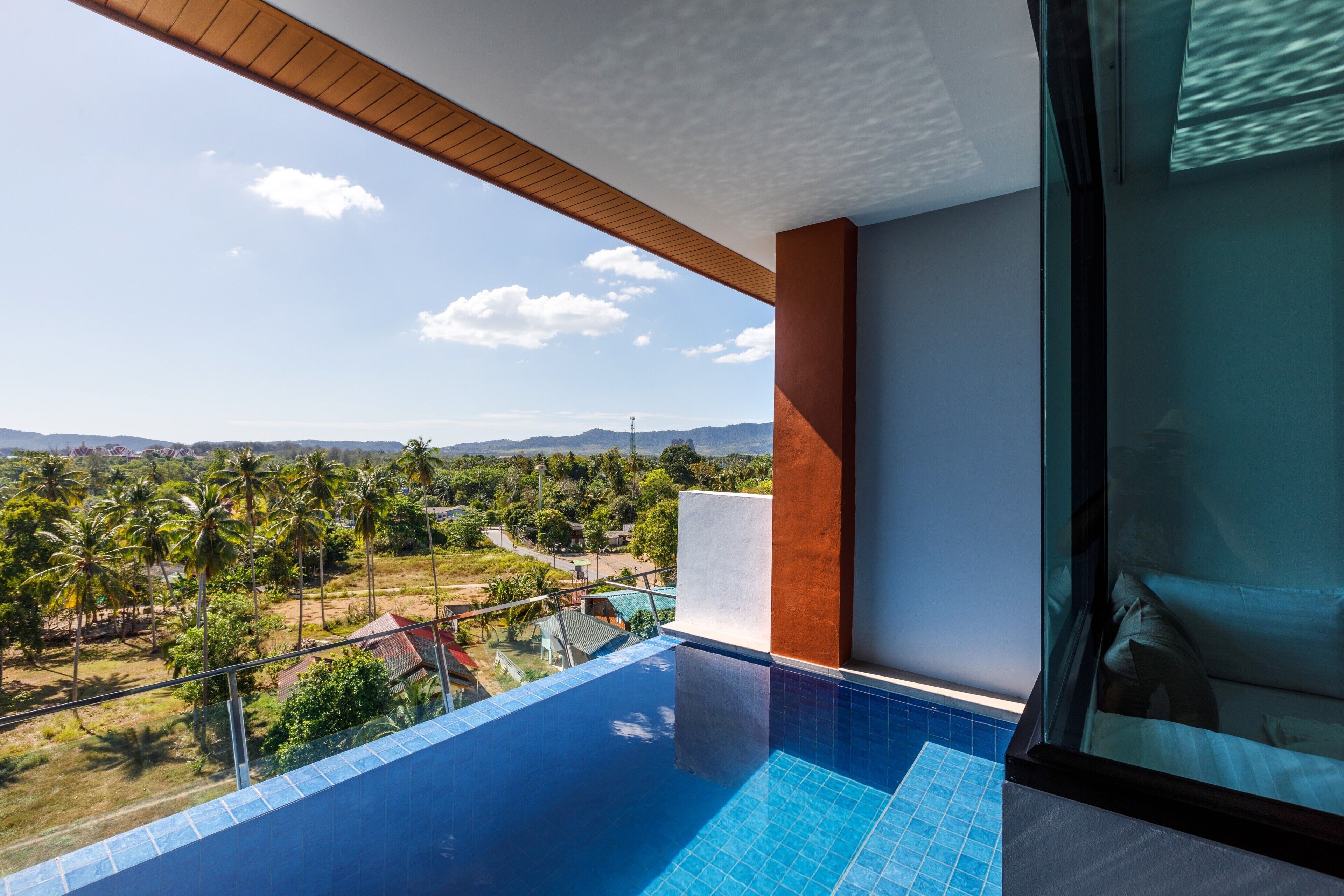 A modern hotel room with a large balcony, a swimming pool, and a view of palm trees and mountains.