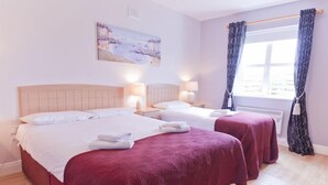 A hotel room with two beds, each with a red bedspread and white pillows, a painting on the wall, and a window with curtains. - Upton Court Holiday Cottages (Kilmuckridge)