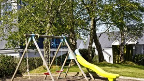 A playground with a slide and swings, surrounded by trees and a fence. - Upton Court Holiday Cottages (Kilmuckridge)