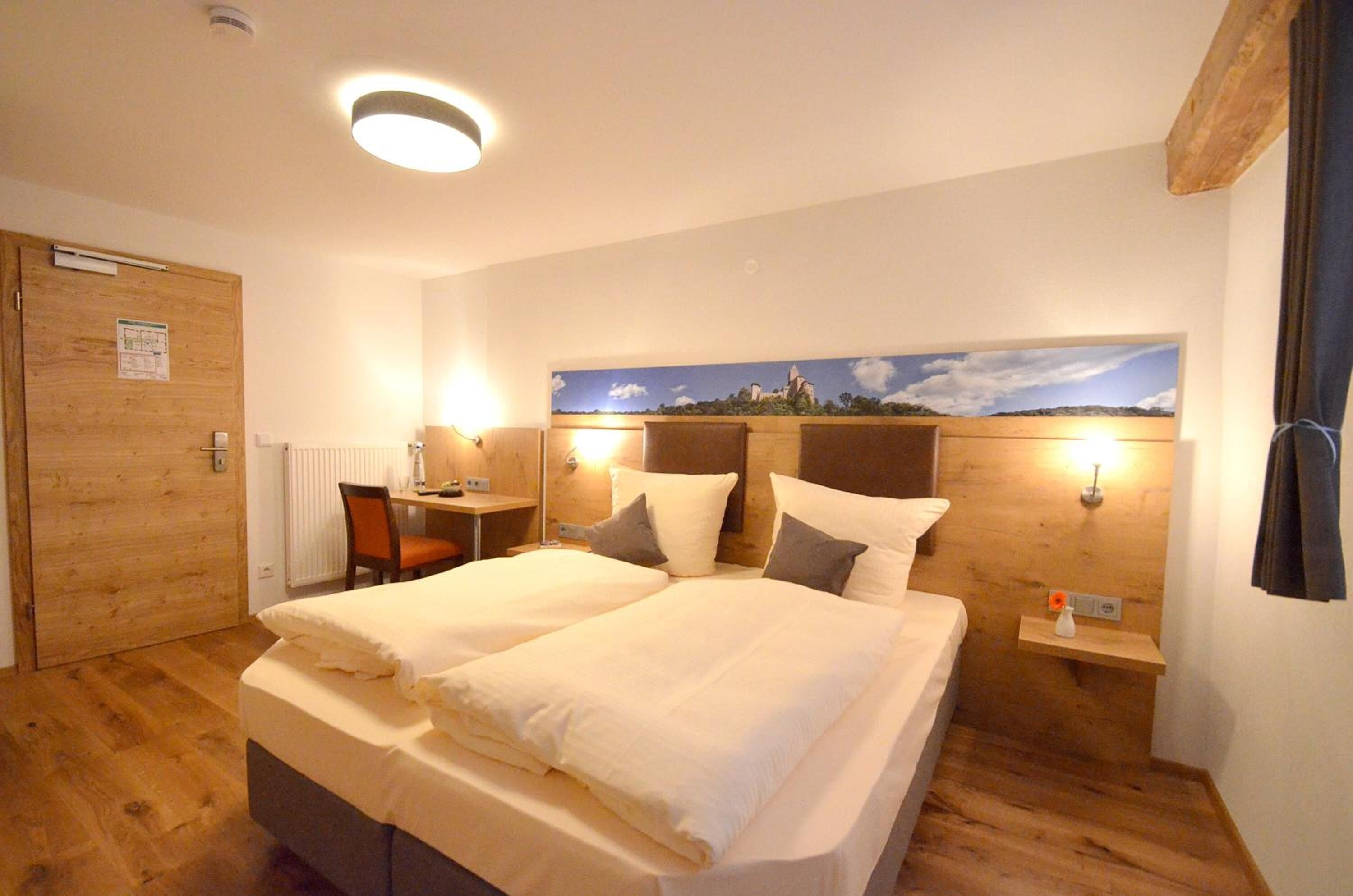 A double bed with two pillows, a wooden headboard, a nightstand with a lamp, a desk with a chair, and a painting on the wall.