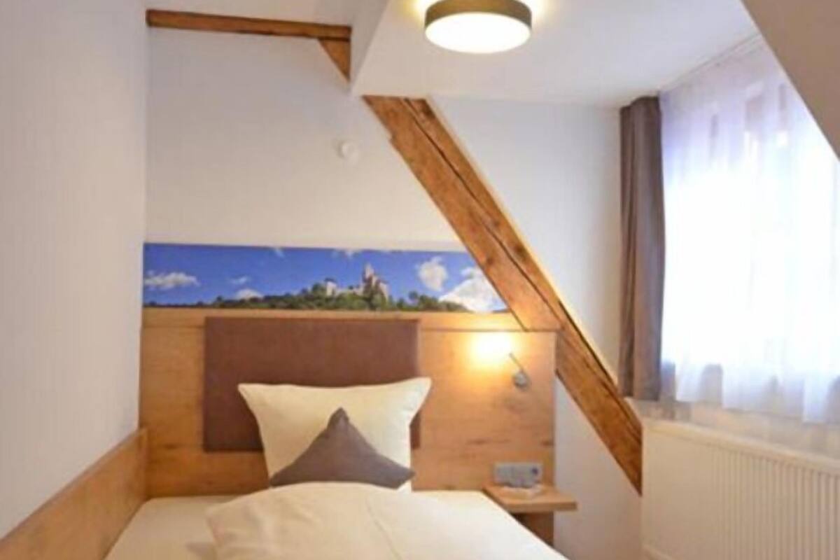 A bedroom with a wooden headboard, a bed with white and brown bedding, a nightstand, a wall-mounted lamp, and a painting above the bed.