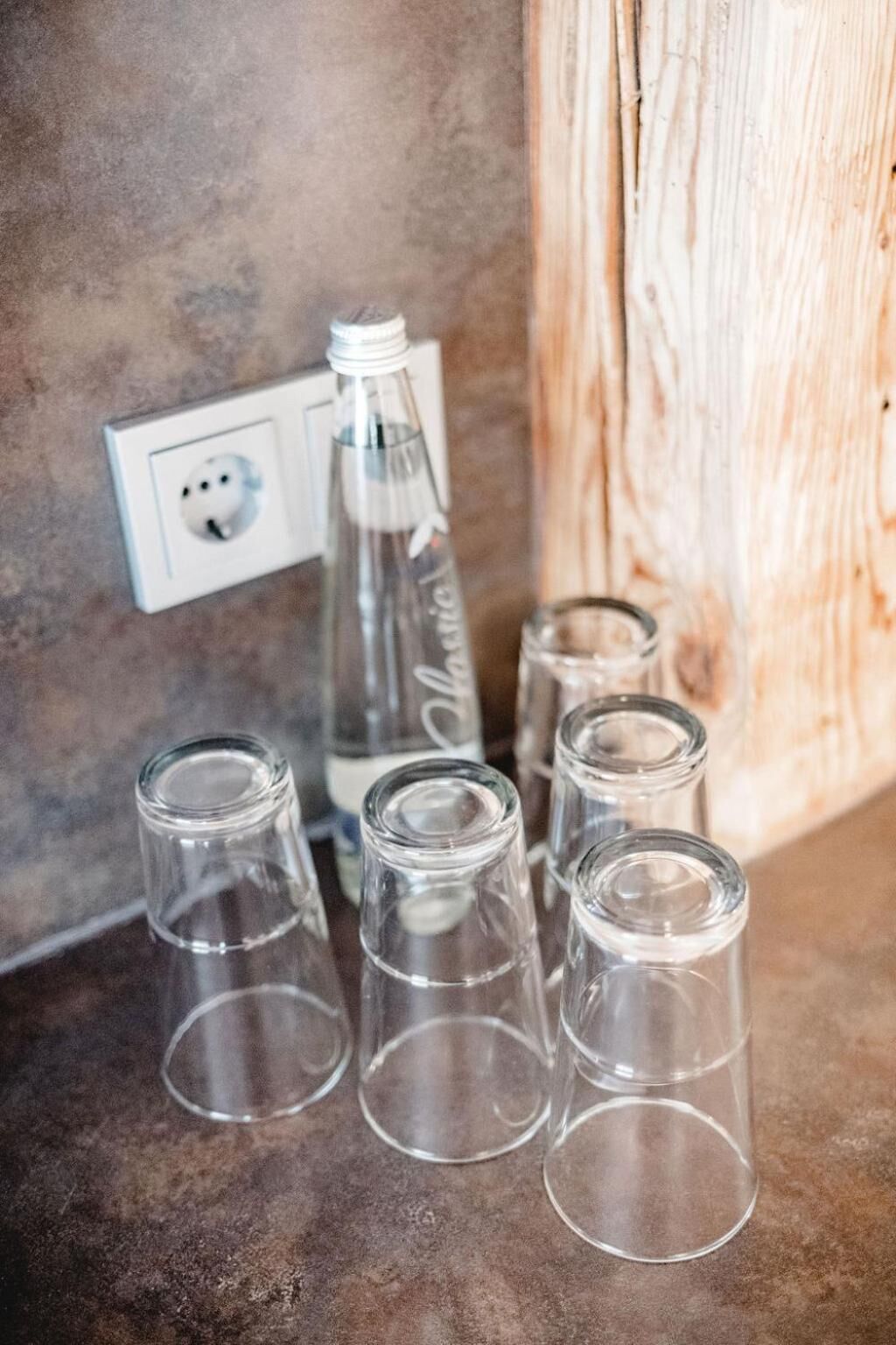 A set of clear glass bottles and glasses on a textured surface near a wooden structure and a white electrical outlet.