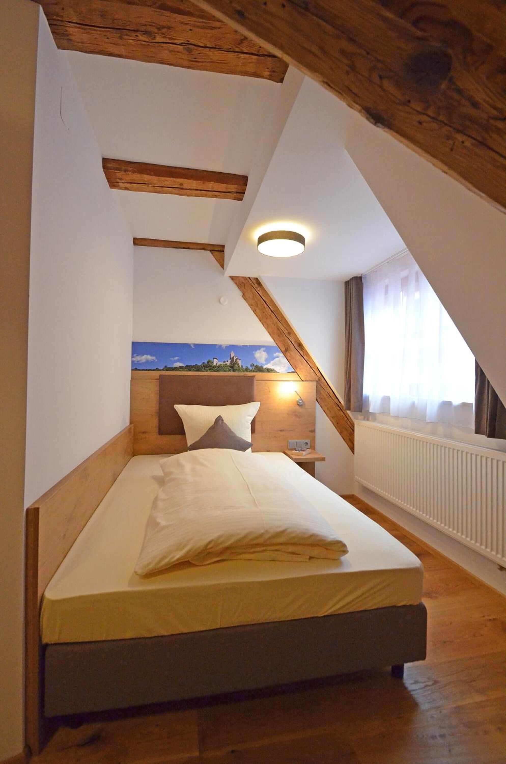 Et soveværelse med skråvæg, træbjælker, en seng med hvide sengetøj, et natbord og en lampe monteret på væggen.
