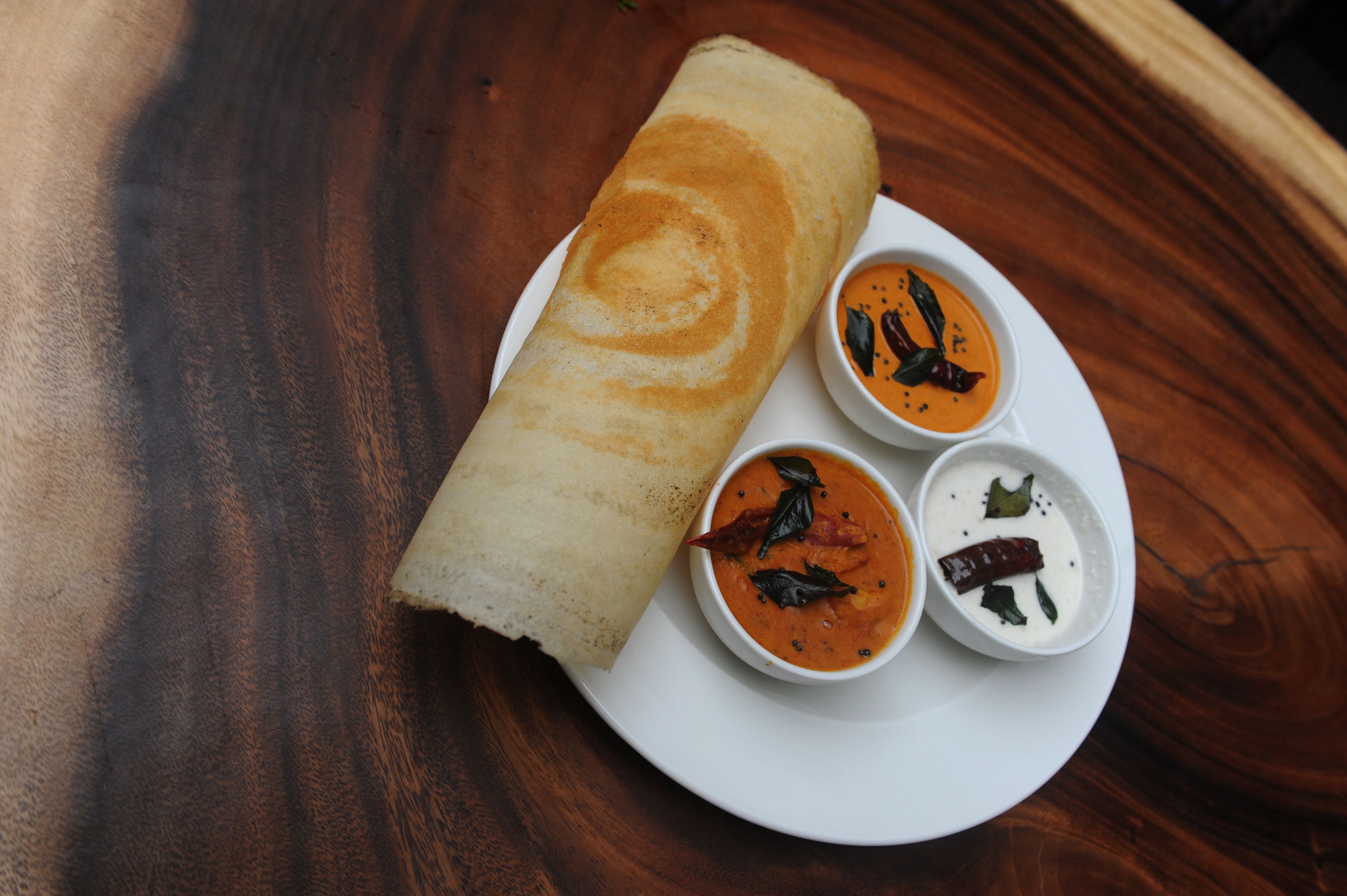 A plate with a rolled-up dosa, two small bowls of chutney, and a white plate on a wooden table.