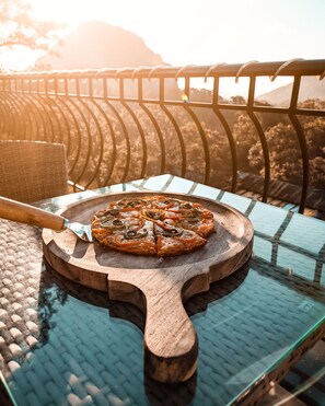 Una pizza sobre una tabla de madera en una mesa de vidrio, con una barandilla metálica al fondo.