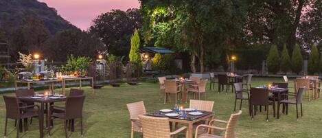 An outdoor dining area with tables and chairs, surrounded by trees and a view of hills.