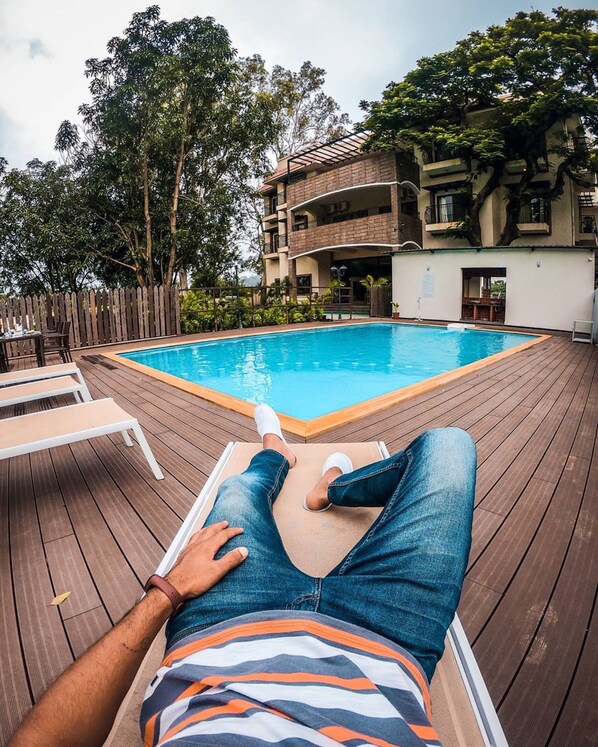 A person relaxing by a pool with a wooden deck, surrounded by a fence and trees. - The Summer House, Pachmarhi - AM Hotel Kollection (Pipariya)