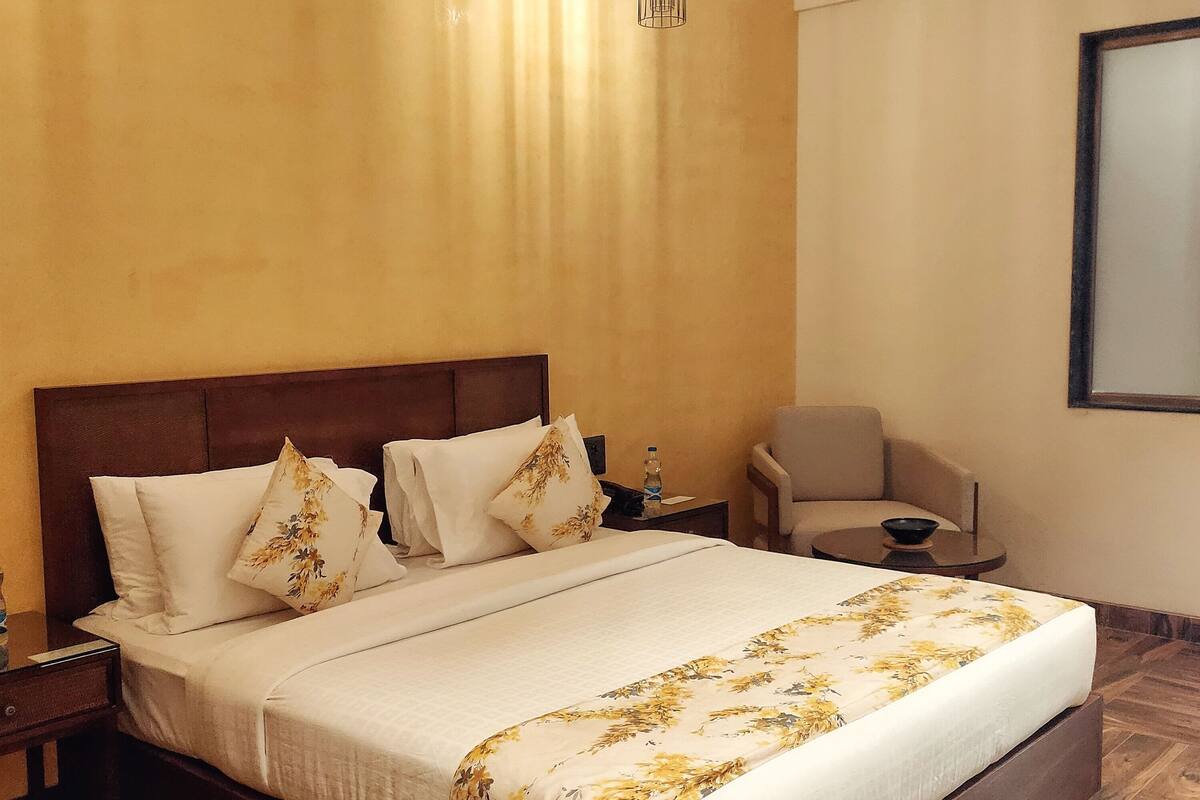 A large bed with a wooden headboard, a bedside table with a water bottle, and a chair in a room with wooden flooring and a large window.