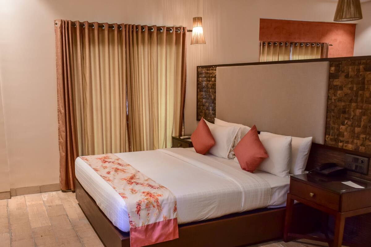 A hotel room with a large bed, a wooden nightstand, a desk, and a wall-mounted lamp.