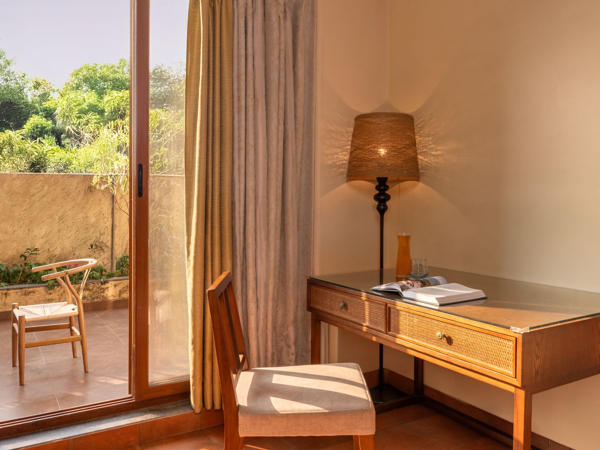 A room with a wooden desk, a lamp, a chair, and a view of greenery outside.
