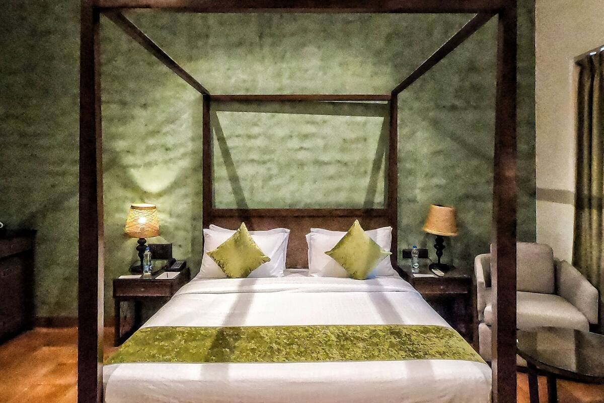A four-poster bed with a green canopy, two bedside tables with lamps, a chair, and a wooden floor.