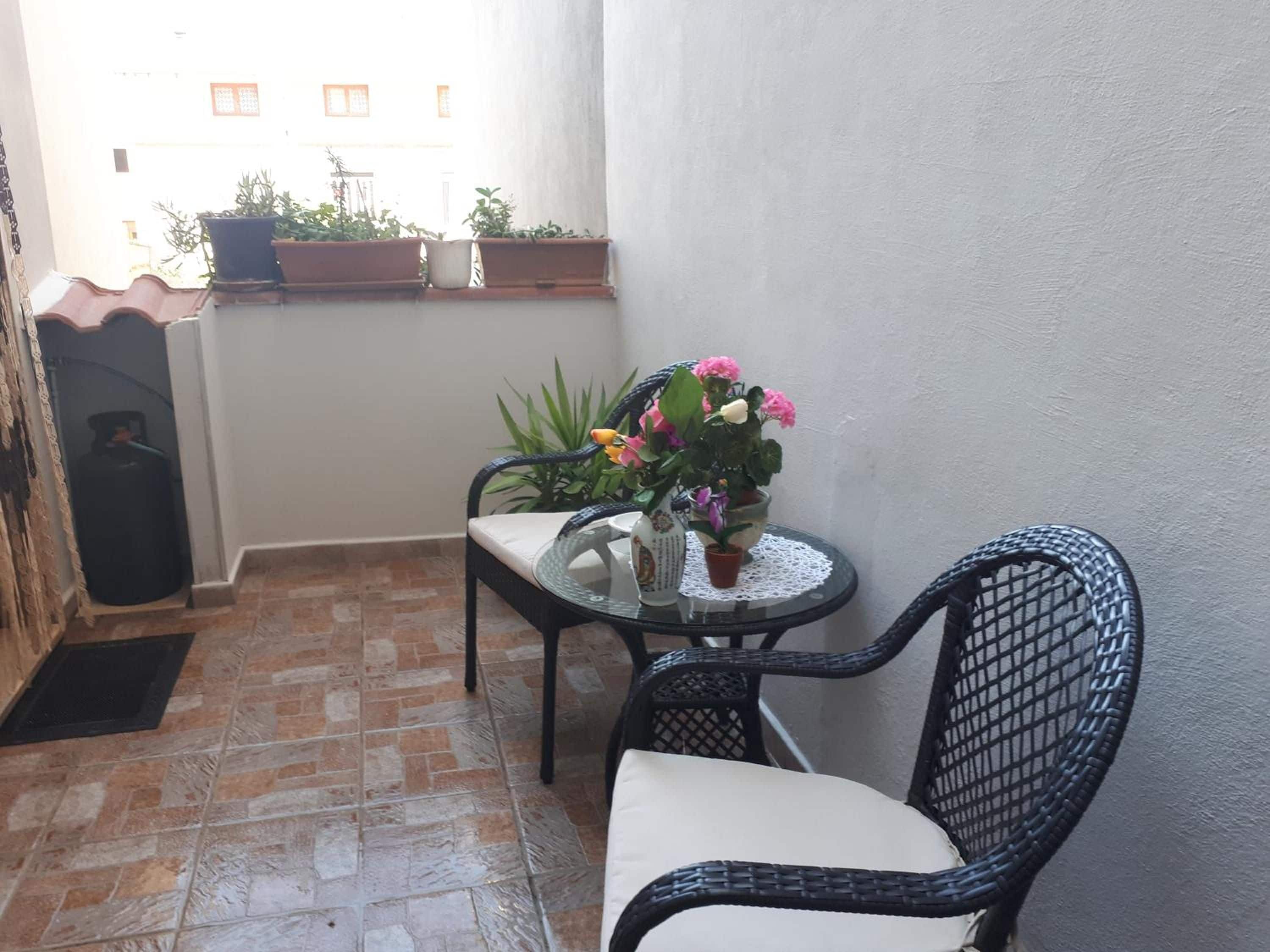Un pequeño balcón con suelo de baldosas, una mesa con un florero, una silla y plantas en macetas.