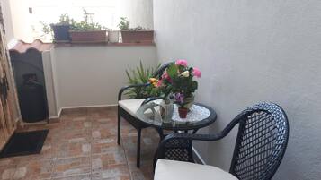 Un pequeño balcón con suelo de baldosas, una mesa con un florero, una silla y plantas en macetas.