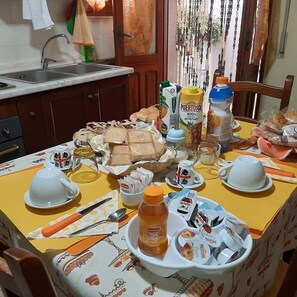 A dining table with a variety of food items, including bread, pastries, and condiments, set in a kitchen with wooden cabinets and a window with curtains. - B&B Da Andreina (Guspini)