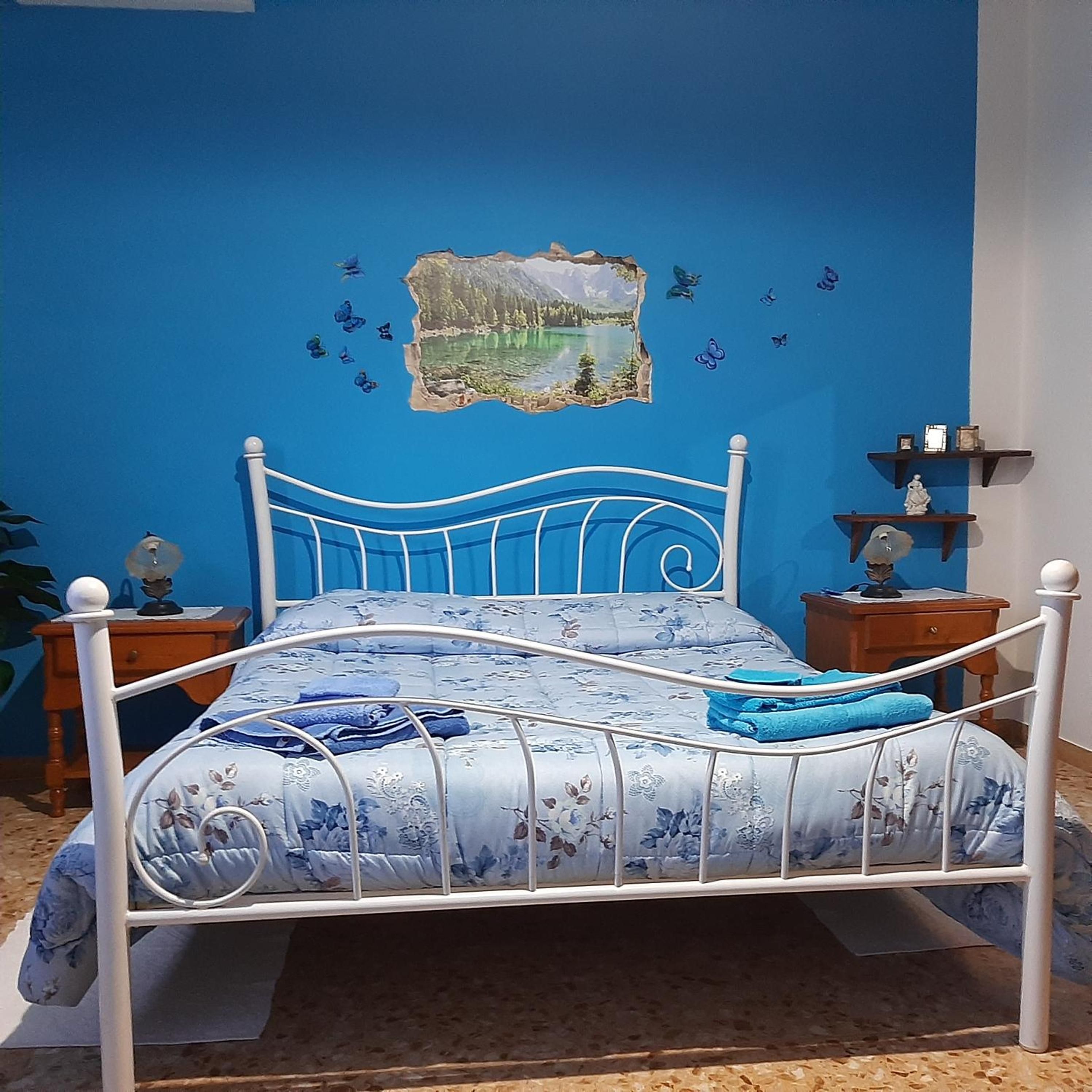Un dormitorio con cama, mesita de noche, una pared con un cuadro y estantería con objetos.