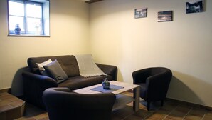 A living room with a brown sofa, a small table, and a black armchair. There is a window with a vase on the windowsill and three framed pictures on the wall. - Appartments Tilia (Quedlinburg)