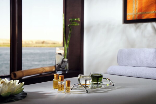 A spa setting with a lotus flower, bottles of essential oils, and a tray with a glass of water and a green drink, viewed through a window overlooking a body of water.