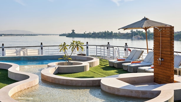Un espace piscine avec vue sur l’eau, des chaises longues et une structure en bois surmontée d’un auvent.