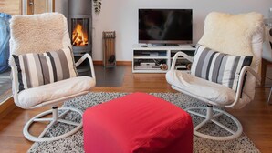Ein Wohnzimmer mit zwei Sesseln, einem roten Hocker, einem Fernseher und einem Kamin.