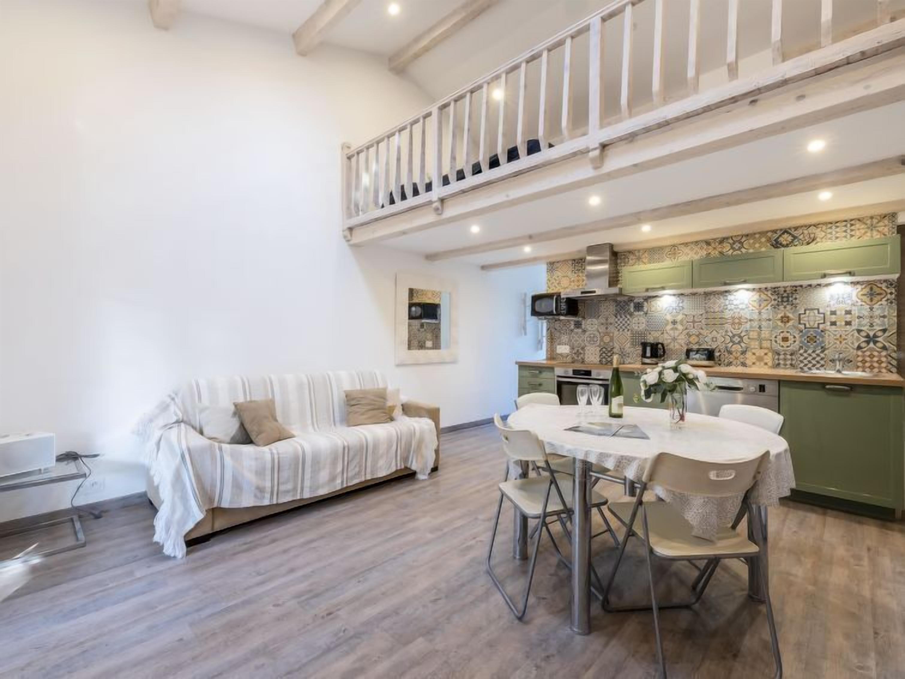 Ein modernes Wohnzimmer mit einer Couch, einem Esstisch und Stühlen. Im Hintergrund ist eine Küche mit gemusterten Fliesen und grünen Schränken zu sehen.