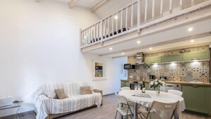 Un salon moderne avec un canapé, une table à manger et des chaises. On aperçoit, au fond, une cuisine avec des carreaux décoratifs et des meubles de couleur verte.