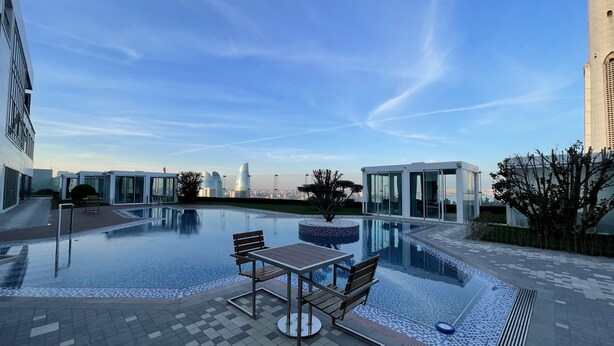 Un área de piscina en la azotea con mesa y sillas, rodeada de edificios modernos y un cielo despejado.