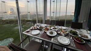 Una mesa de comedor dispuesta con platos, cubiertos y vasos, con vistas a un paisaje urbano y una masa de agua.