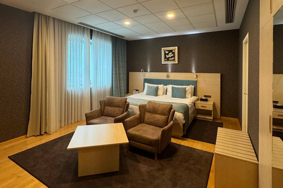 Une chambre d’hôtel moderne, dotée d’un grand lit, de deux fauteuils, d’une petite table et d’une grande fenêtre avec des rideaux.