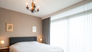 Une chambre d’hôtel avec un grand lit, un banc en bois, un lustre et deux tableaux représentant des motifs floraux, encadrés et accrochés au mur.