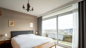 Une chambre d’hôtel moderne avec un grand lit, un banc en bois, un lustre et une vue sur la ville à travers de grandes fenêtres.
