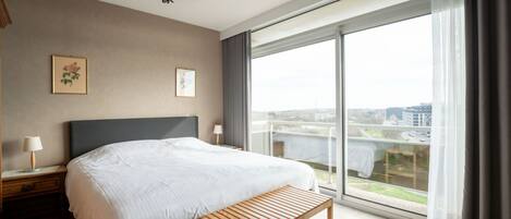 A modern hotel room with a large bed, a wooden bench, a chandelier, and a city view through large windows.